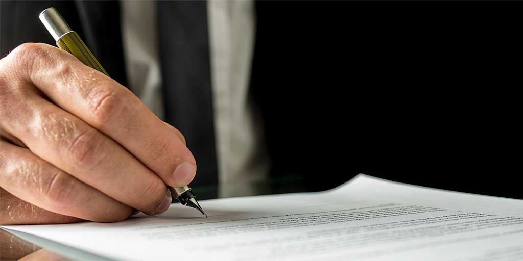 A consultant wearing a black suit signs an expert witness report with a pen on a table. The hand is in focus, highlighting the act of writing, while the background is slightly blurred.